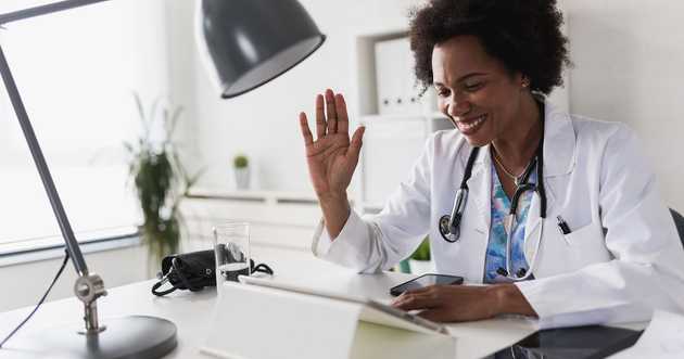 woman doctor at desk waving at tablet